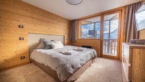 Quarto com parede de madeira, cama com almofadas, mesa de cabeceira e janela com vista para montanhas.