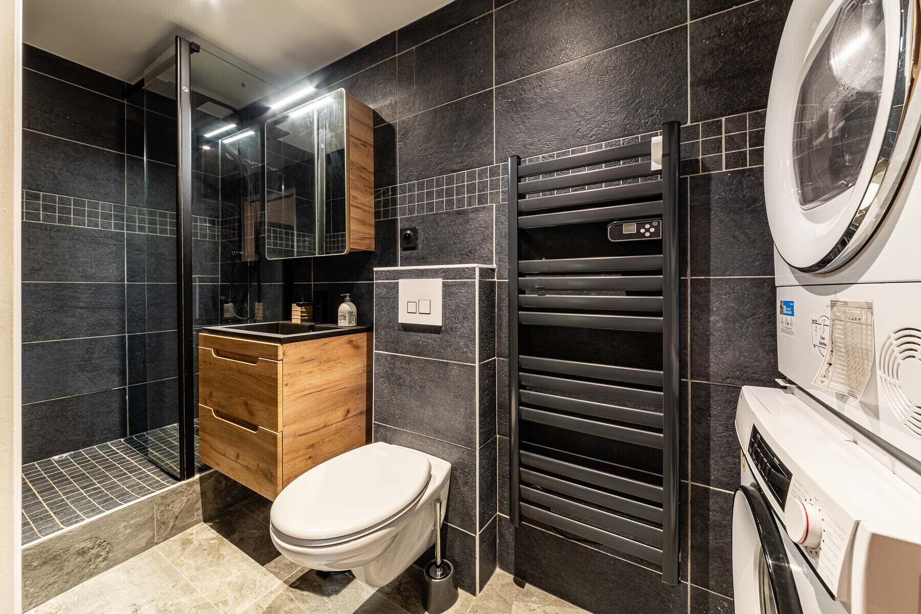 A modern bathroom with a toilet, wooden vanity, black tiled walls, a glass shower enclosure, and a built-in washing machine.