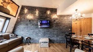 Een gezellige woonkamer met een stenen muur, een daaraan bevestigde televisie, een houten eettafel met stoelen en een bank met hartvormige kussens.