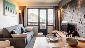 Una habitación de hotel moderna con un sofá gris, una mesa de comedor con sillas, un televisor y un amplio ventanal con vistas a la montaña.