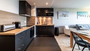 Una cocina moderna con armarios oscuros, encimera de madera y horno empotrado. Hay una zona de comedor con mesa y sillas, y un sofá al fondo.