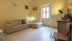 Una sala de estar con un sofá seccional color beige, una silla de madera, un escritorio con una lámpara y una ventana.