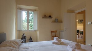Una habitación de hotel con cama, ventana, silla y una mesita con lámpara.