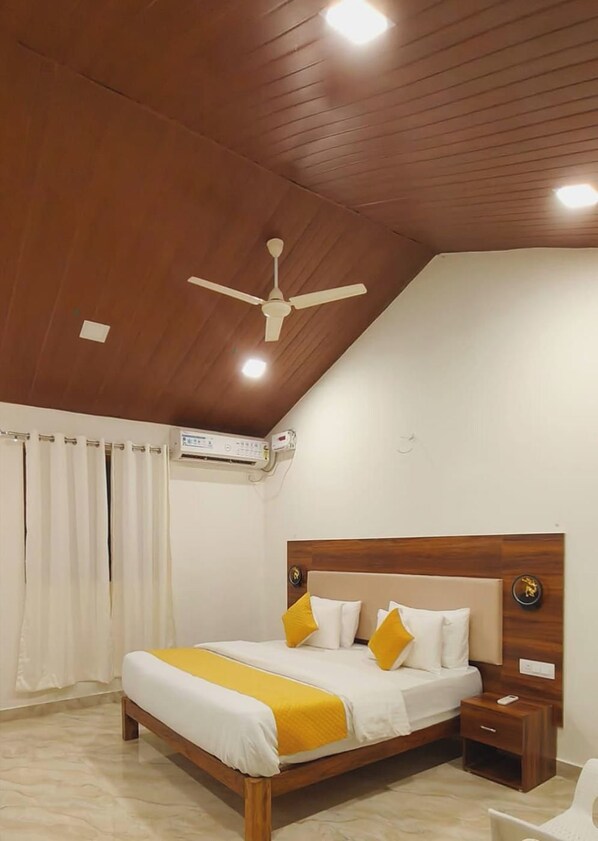A bedroom with a wooden ceiling, a ceiling fan, an air conditioning unit, a bed with white and yellow bedding, and a bedside table with a lamp. - Gondwana Jungle Resort (Chimur)