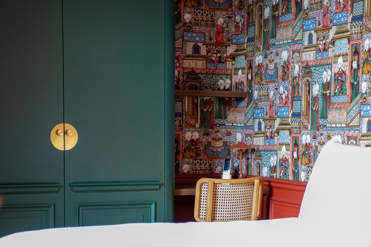 Una habitación con puerta color turquesa, papel tapiz estampado y una silla de mimbre.