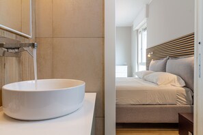 A modern bathroom with a large white vessel sink and a wooden headboard in the adjacent bedroom.