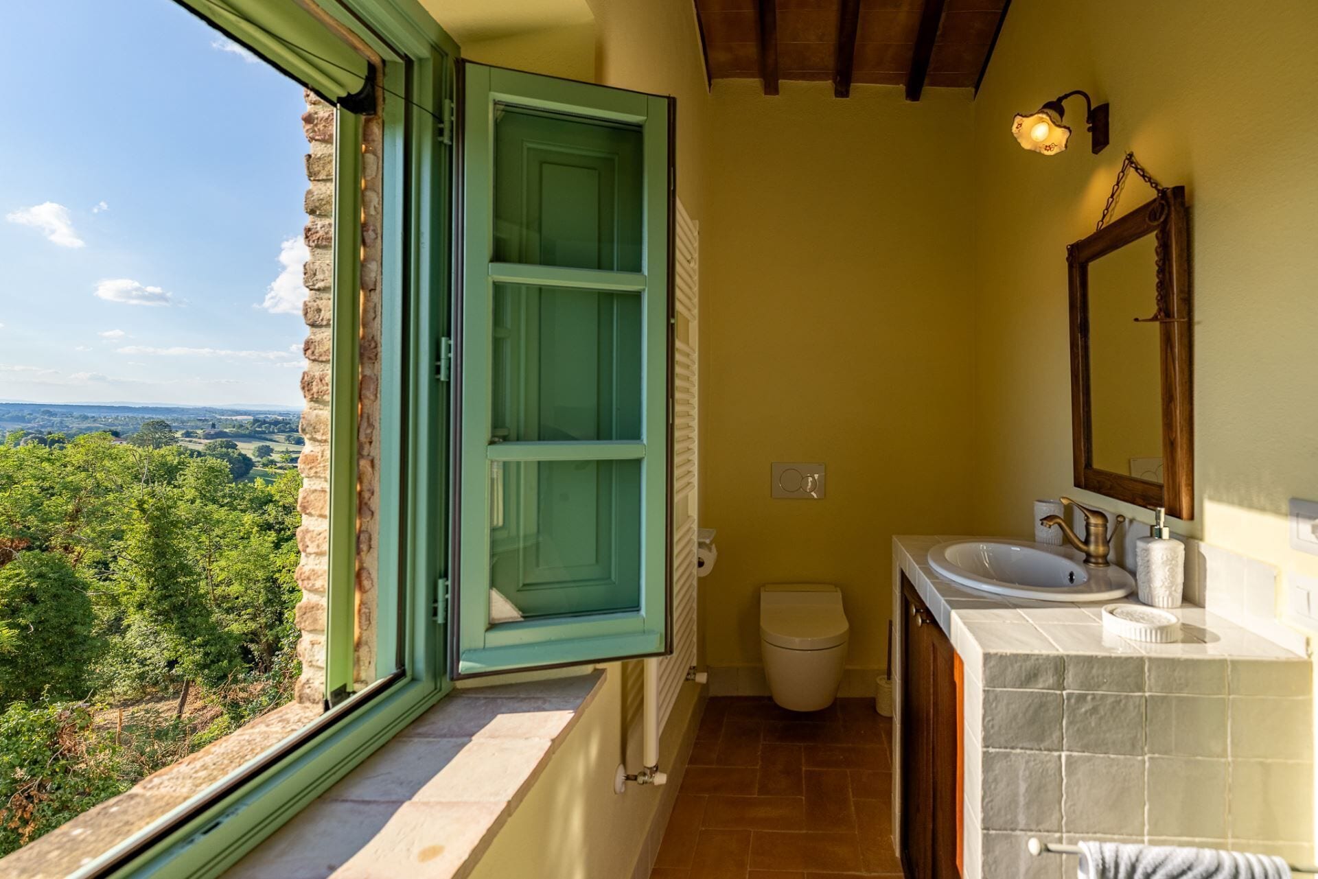 Une salle de bain avec vue sur un paysage verdoyant, des toilettes, un lavabo avec un miroir et un porte-serviettes.