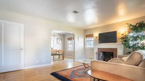 A living room with a sofa, a TV, a fireplace, and a dining area in the background. - Chic Boho Bungalow in Land Park! (Sacramento)