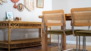 A dining area with two wooden chairs with woven backs and green cushions, a wooden table, and a wicker side table with a picture frame and decorative items on the wall. - Chic Boho Bungalow in Land Park! (Sacramento)
