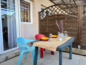 Een buiten eethoek met een tafel gedekt voor twee personen, met een tafelkleed met bloemenmotieven en een vaas met gedroogde bloemen. Er zijn twee stoelen, één blauw en één rood, en een houten poort op de achtergrond.