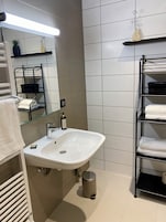 Un baño con un lavamanos blanco, un espejo, un perchero para toallas, un estante con un florero y un cubo de basura.