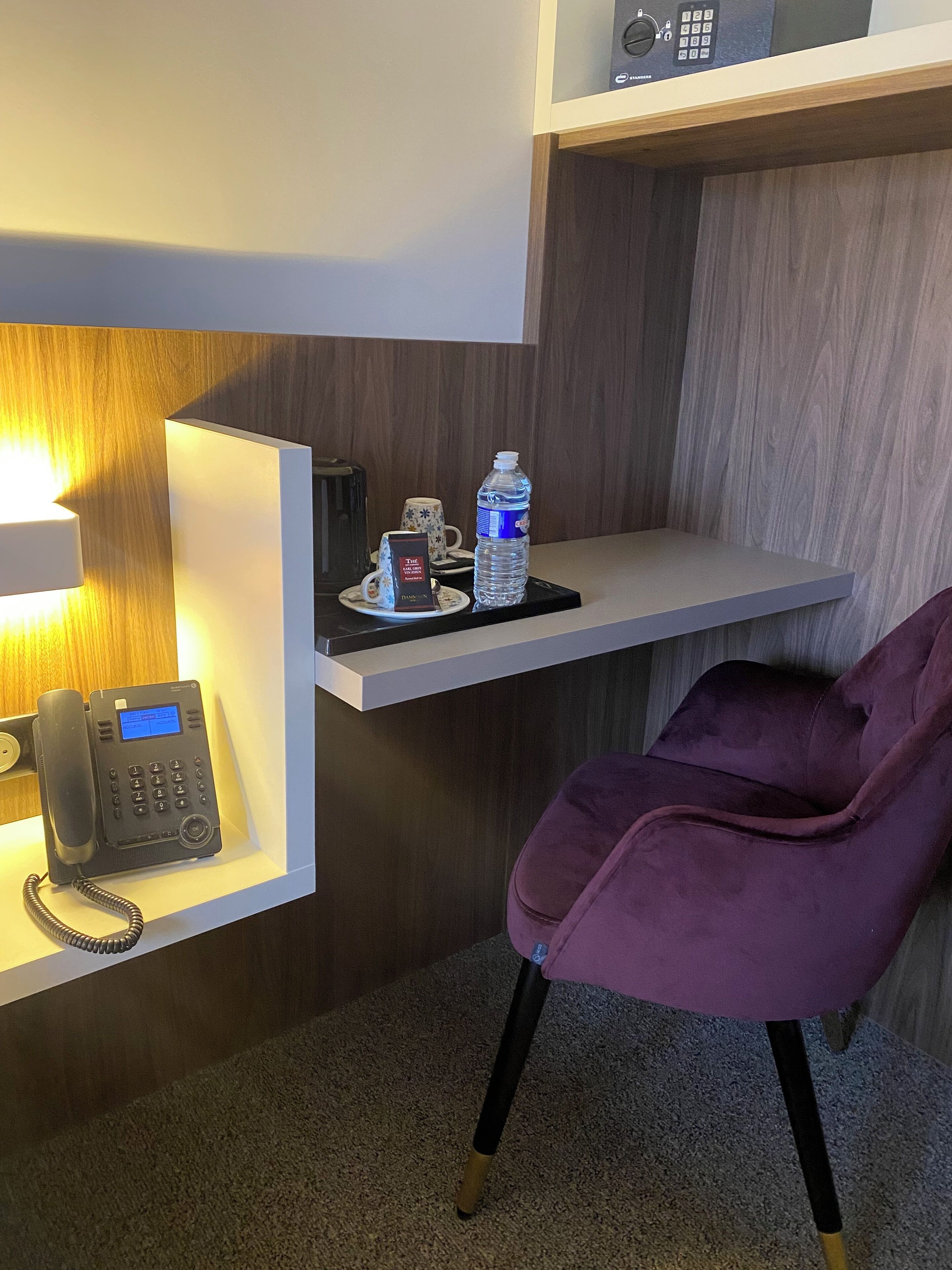 Zona de recepción de hotel con un teléfono moderno, una botella de agua, una taza y una silla morada.