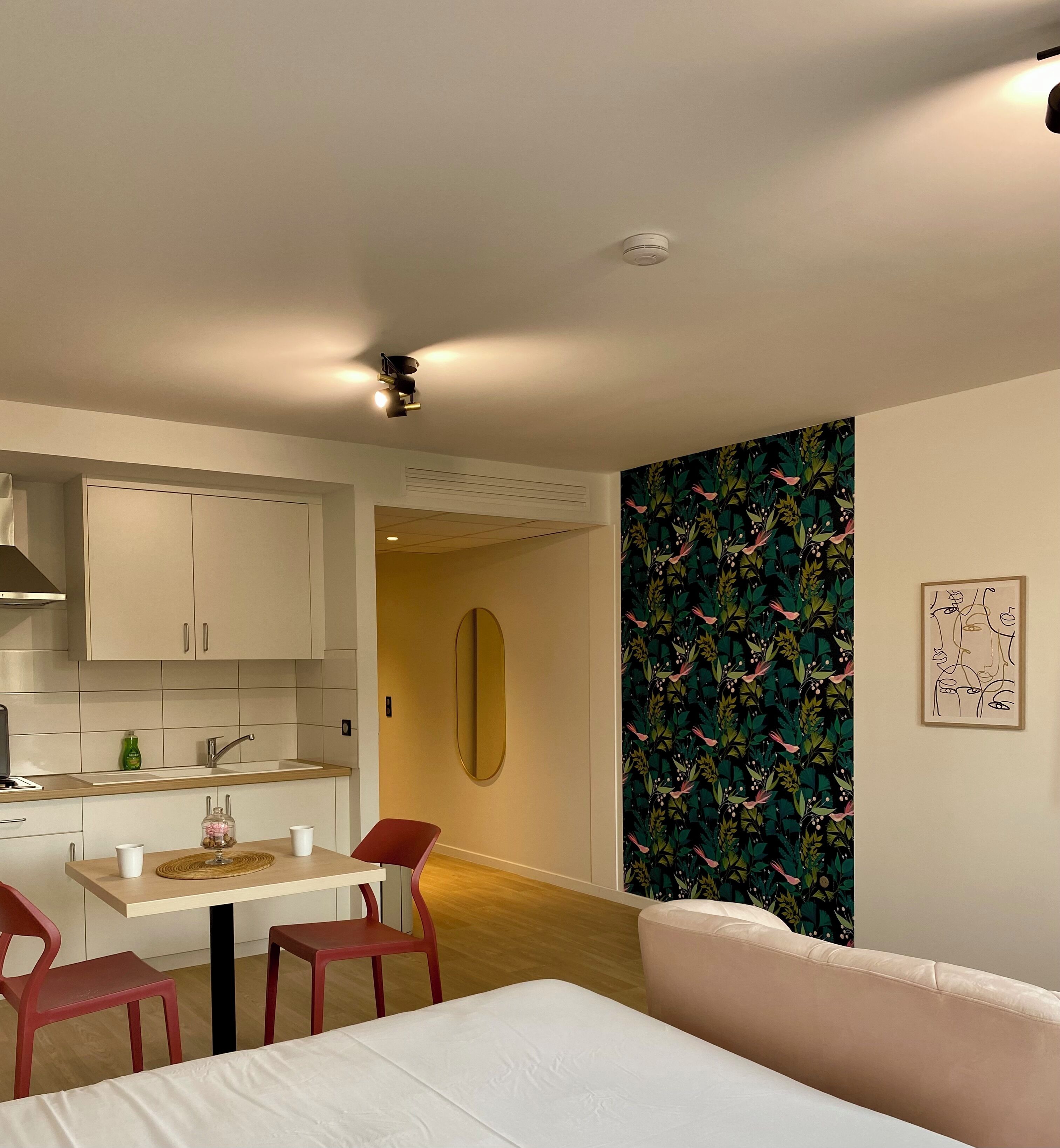 A modern living room with a sofa, a dining table with chairs, a kitchen area with cabinets, and a wall with a floral pattern.