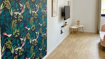 A modern living room with a floral wallpaper, a flat-screen TV, a small round table with a lamp, and a chair.