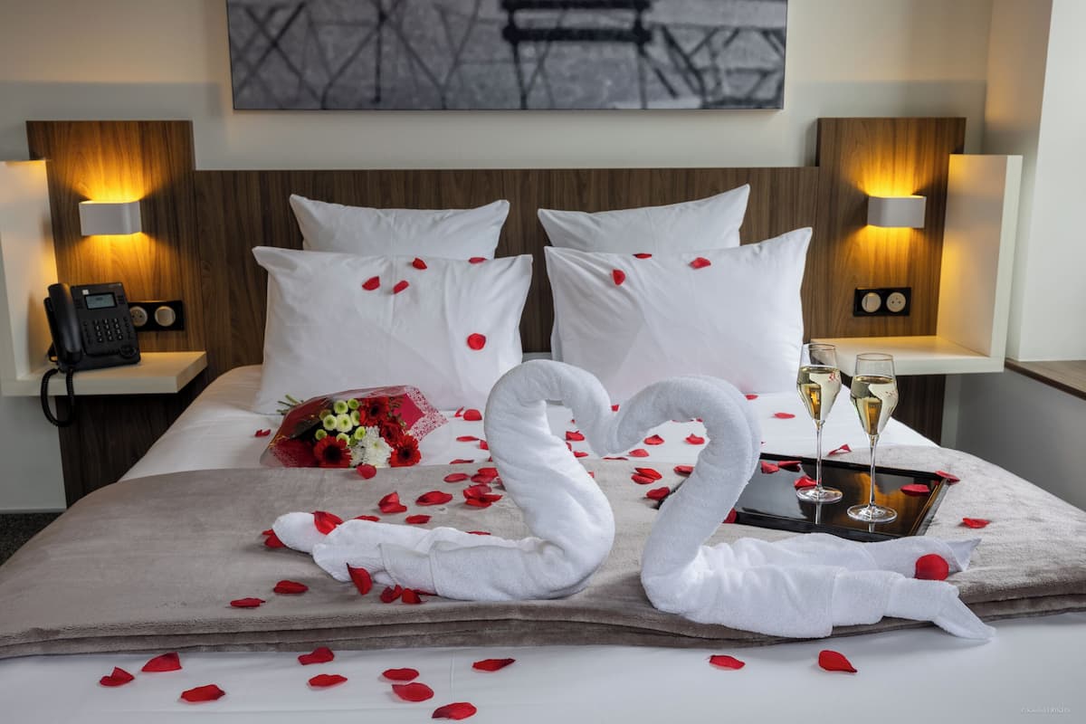 Habitación de hotel con cama, almohadas, un ramo de flores, pétalos de rosa, dos toallas con forma de cisne y dos copas de champán.