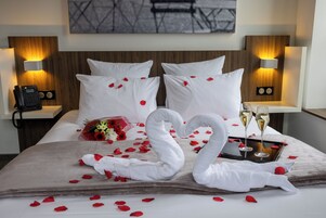A hotel room with a bed, pillows, a bouquet of flowers, rose petals, two swan-shaped towels, and two champagne glasses.
