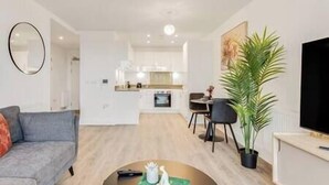 A modern living room with a grey sofa, a coffee table, and a dining area with a table and chairs. There is a kitchen in the background with white cabinets and a stove. A large potted plant is on the right side. - CannyCity 1 Bedroom APT (London)