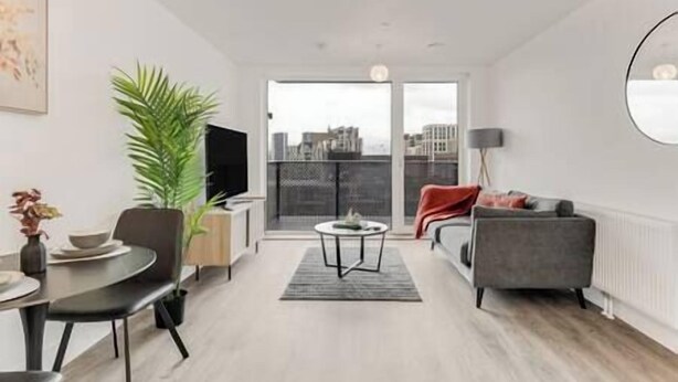 A modern living room with a sofa, a dining table, and a TV. There is a large window with a view of buildings. - CannyCity 1 Bedroom APT (London)