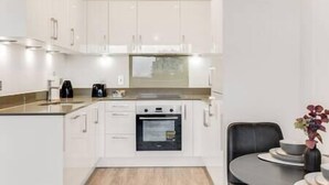 A modern kitchen with white cabinets, a built-in oven, and a small dining area with a table and chairs. - CannyCity 1 Bedroom APT (London)