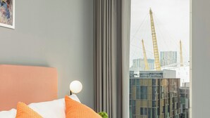 A modern hotel room with a large bed, orange and white pillows, a view of a cityscape with cranes, and a grey curtain. - ShyPen One - Bedroom APT (London)