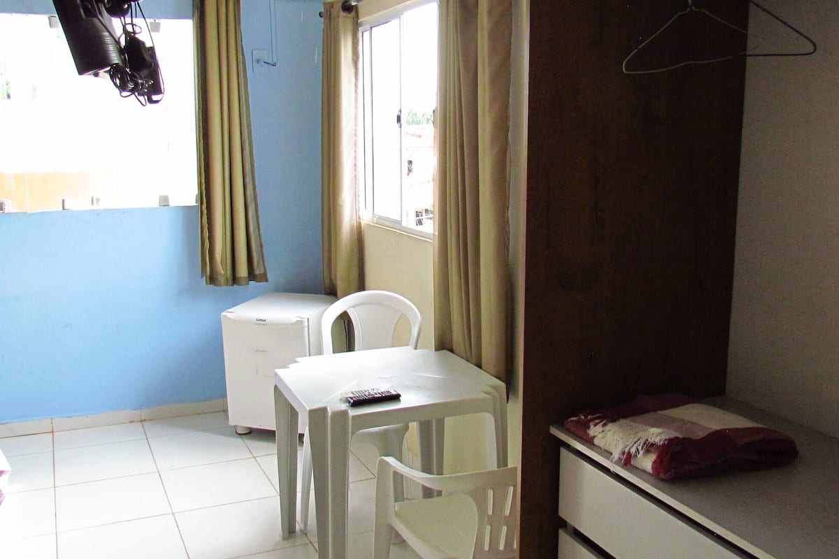 A small, simple room with a bed, a desk, and a chair. There is a window with curtains, a heater, and a hanging coat.