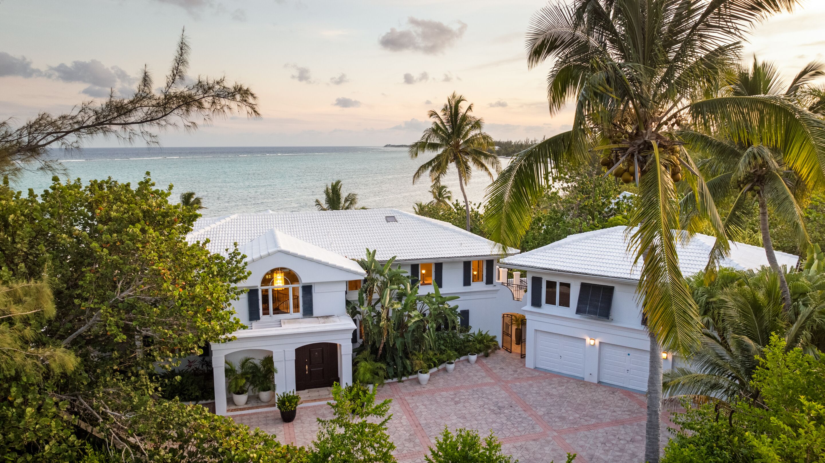A white two-story house with a tiled roof, surrounded by lush greenery and a view of the ocean.