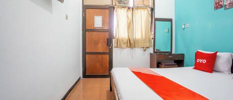 Quarto de hotel com cama, porta de madeira, espelho, mesa de cabeceira e travesseiro vermelho com a palavra 'OYO'.