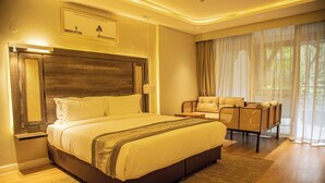A spacious bedroom with a large bed, a wooden headboard, a seating area with a chair and a small table, and a large window with sheer curtains. - Lion Villas (Nairobi)