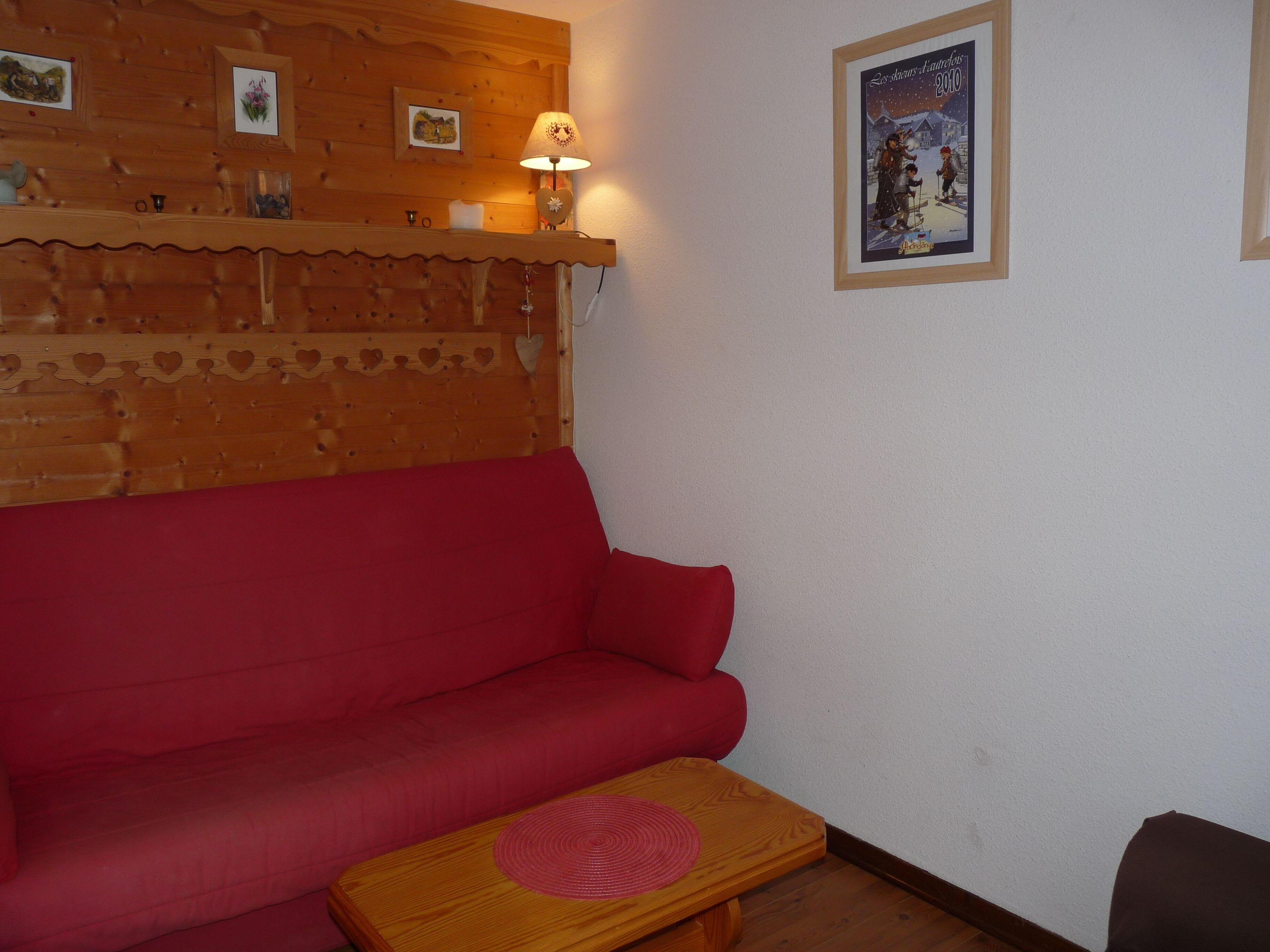 A cozy room with a red sofa, a wooden table with a placemat, and a wall decorated with framed pictures and a lamp.