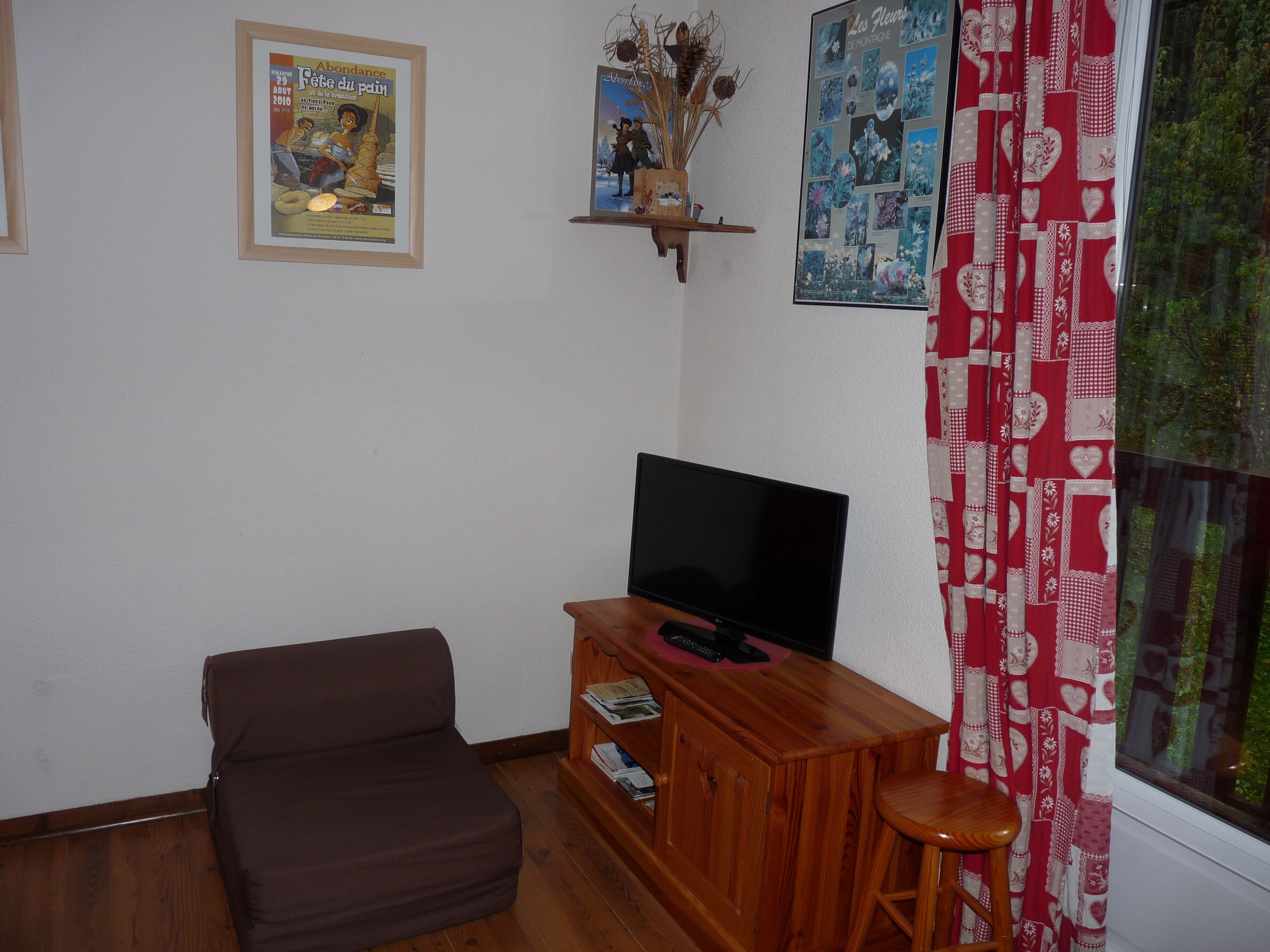 Ein Wohnzimmer mit braunem Sofa, einem TV-Schrank aus Holz mit Flachbildfernseher, einem kleinen Holzschemel und einem Fenster mit roten und weißen Vorhängen.