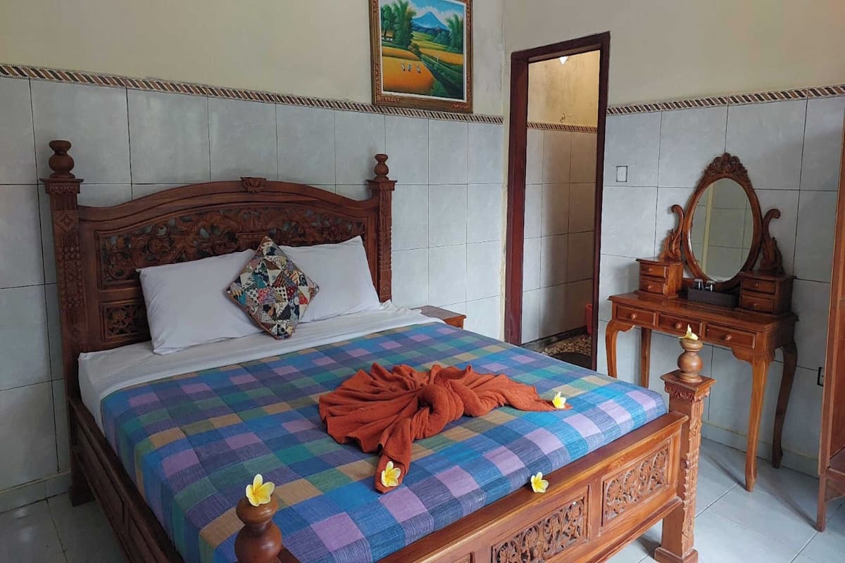 Un lit bien fait, recouvert d’une couverture à carreaux colorés et agrémenté d’une serviette pliée, une commode en bois avec un miroir, et un tableau accroché au mur.