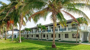 Een hotel met een witte gevel, meerdere balkons en een verzorgd gazon met palmbomen.