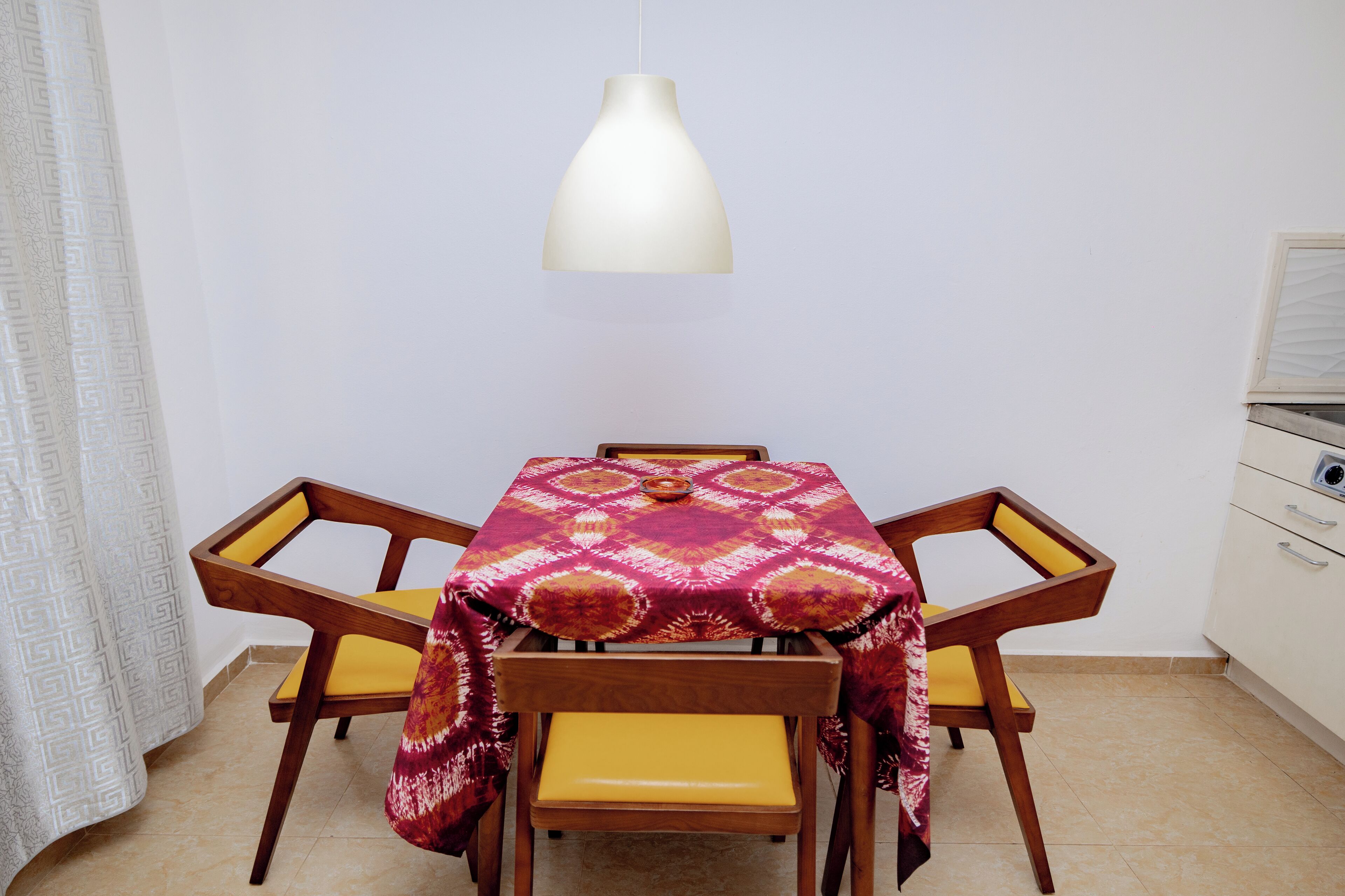 Un comedor con una mesa cubierta por un mantel estampado, sillas de madera con cojines amarillos y una lámpara colgante.