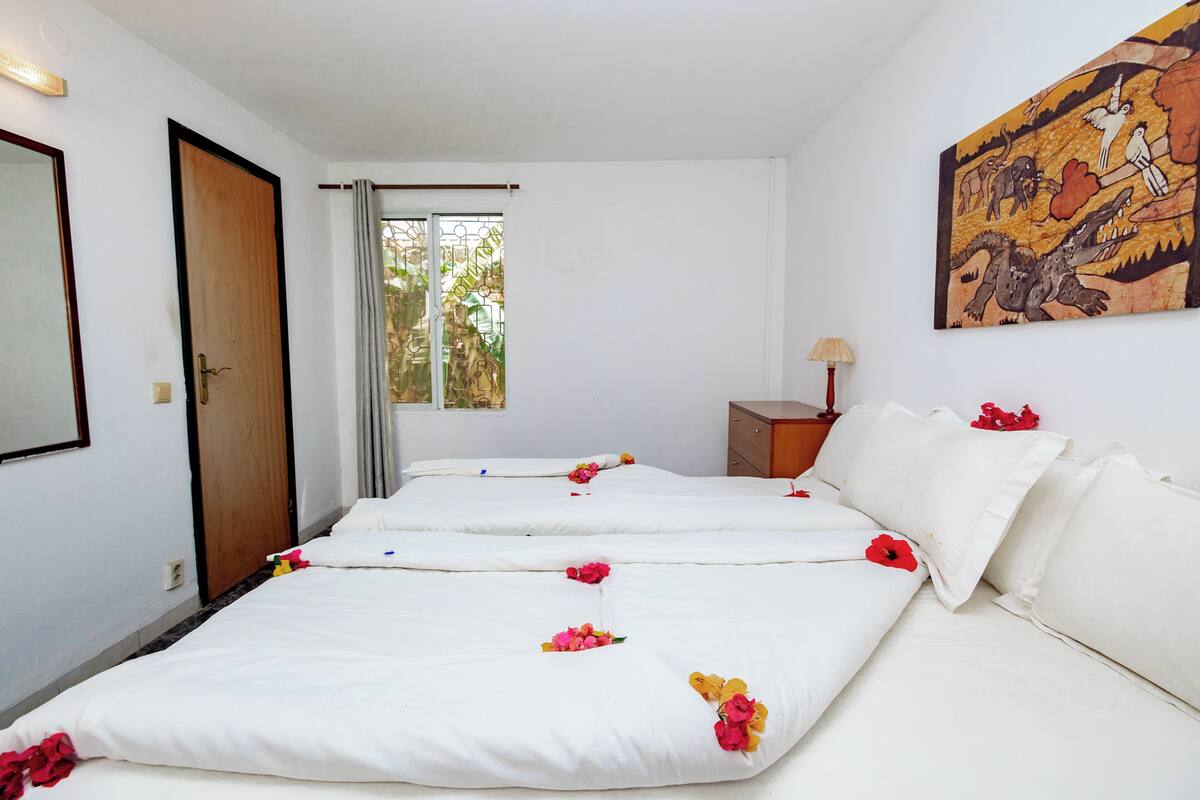 A hotel room with two beds, a wooden door, a window with curtains, a small table with a lamp, and a wall painting.