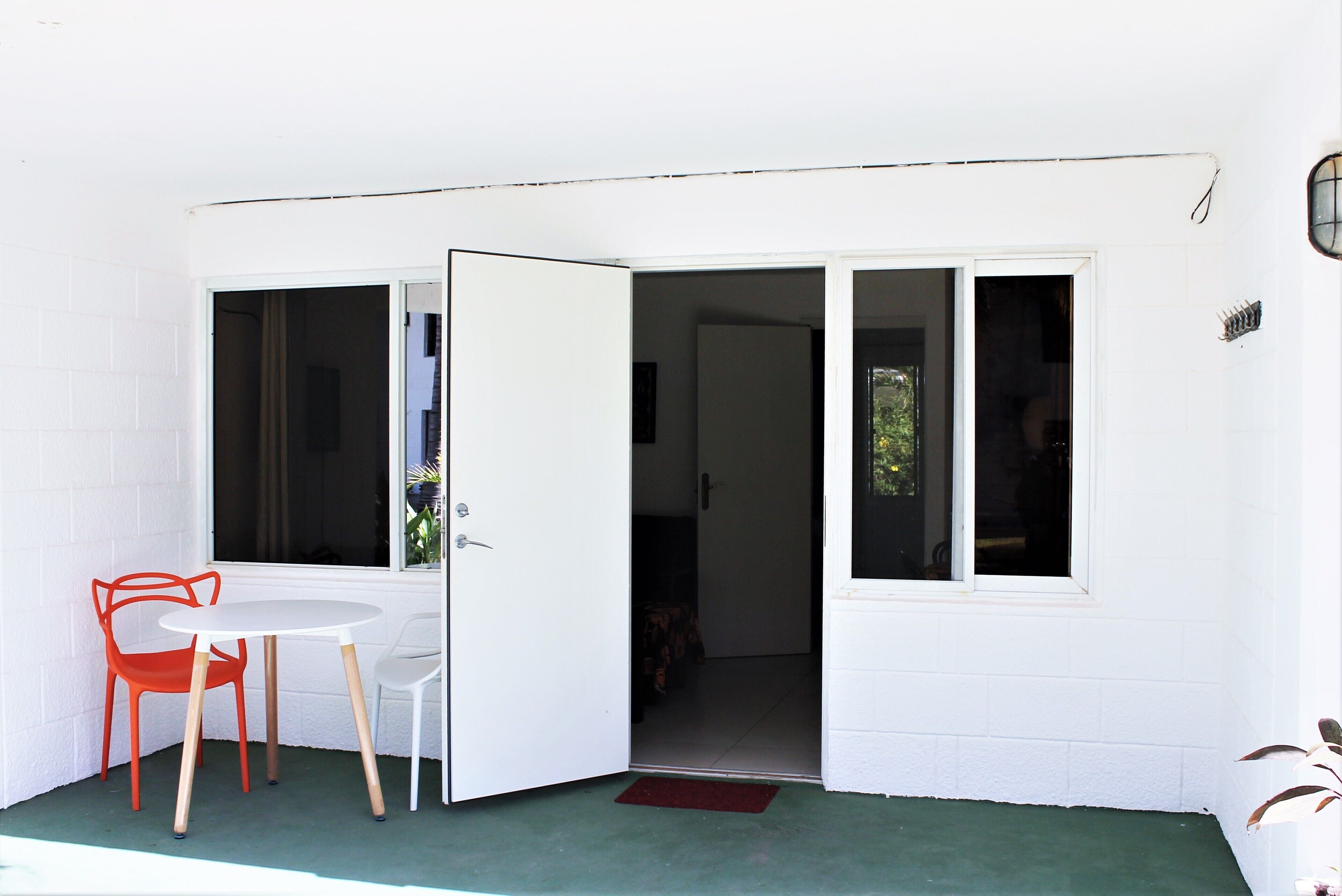 A white exterior wall with a sliding glass door leading to a room. There is a small round table with two chairs, one orange and one white.