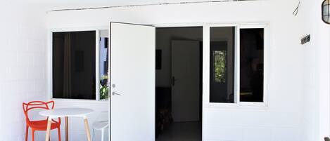 Paredes externas brancas com uma porta de correr de vidro que dá para um quarto. Há uma mesa redonda pequena com duas cadeiras, uma laranja e outra branca.