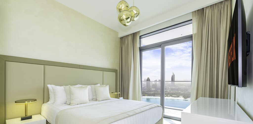 Ein Hotelzimmer mit einem großen Bett, einem Fernseher, einem Schreibtisch und Blick auf eine Stadtlandschaft durch große Fenster.