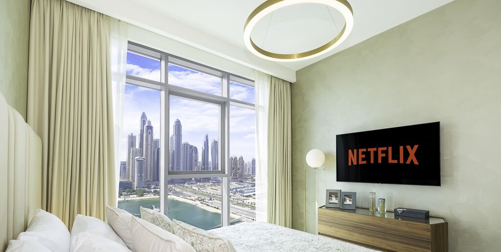 A hotel room with a large window offering a city view, a bed with white linens, a wooden nightstand with a Netflix sign, and a modern light fixture.
