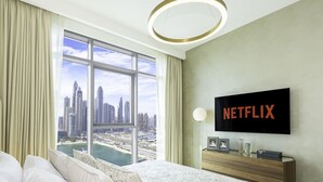 Ein Hotelzimmer mit großem Fenster und Blick auf die Stadt, einem Bett mit weißer Bettwäsche, einem Nachttisch aus Holz mit Netflix-Logo und einer modernen Lampenleuchte.