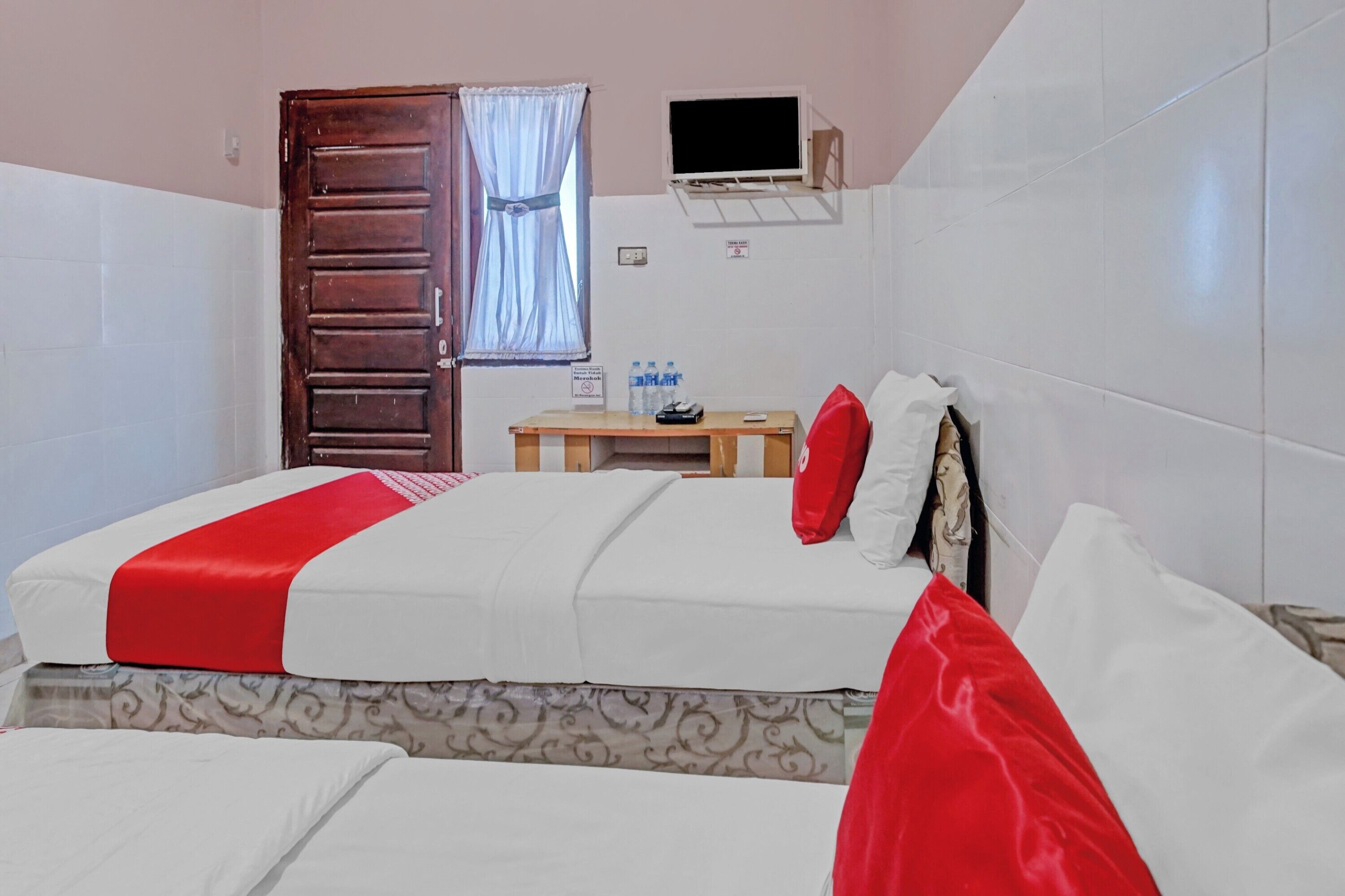 A hotel room with a bed, a small table with a chair, a window with curtains, and a TV mounted on the wall.