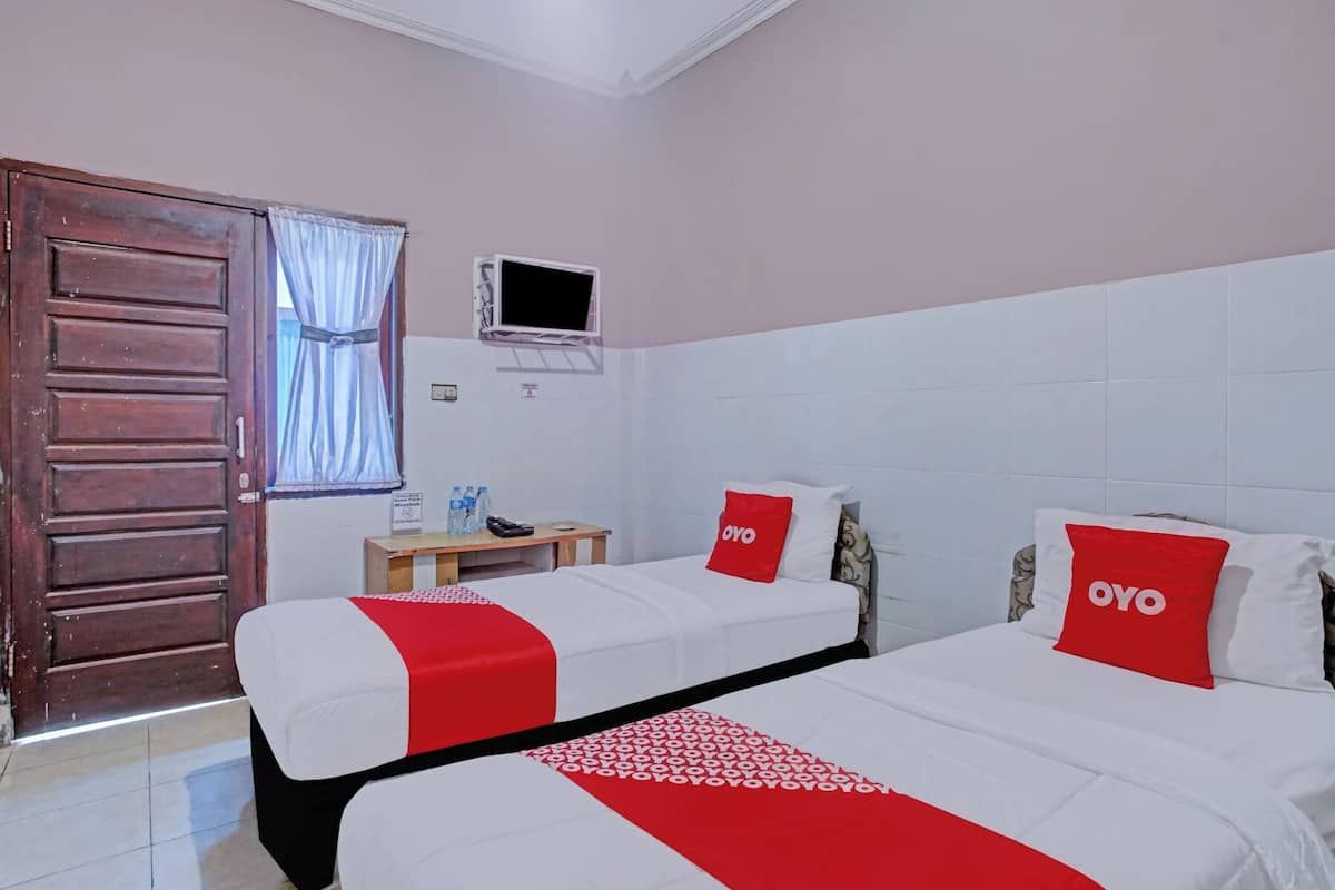 A hotel room with two beds, each with a red and white bedspread and a pillow with "OYO" branding. There is a wooden door, a small table with a water bottle, and a TV mounted on the wall.