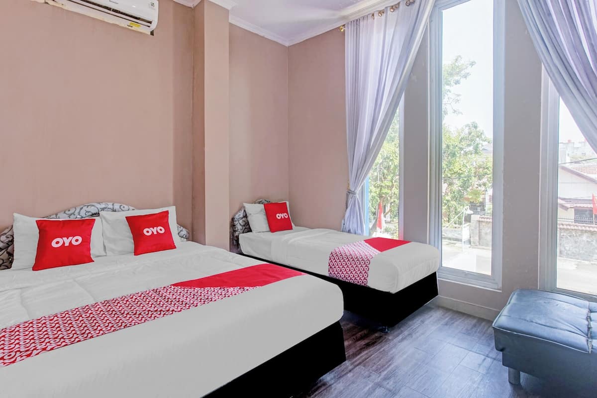 A hotel room with two beds, each with white bedding and red accents, a large window with curtains, and a grey bench.