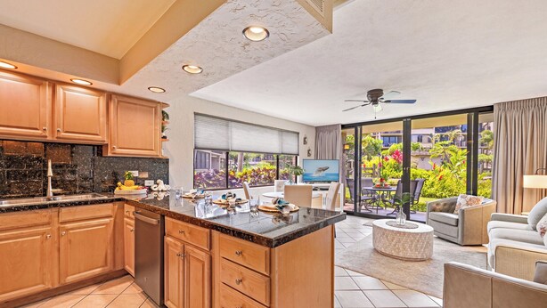 A modern kitchen with wooden cabinets and a large island, a dining area with a TV, and a balcony with a view of a lush garden.