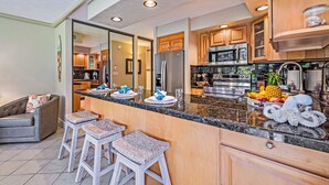 A modern kitchen with granite countertops, stainless steel appliances, and wooden cabinets. There are three bar stools and a dining table set with plates and glasses.