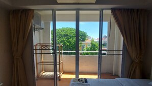 A room with a bed, a window with curtains, an air conditioning unit, and a view of greenery and buildings outside. - Grand Smile Wellness Resort (Pattaya)