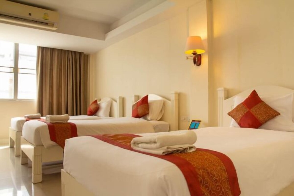 A hotel room with two beds, each with white linens and red accents, a bedside table with a lamp, and a window with curtains.