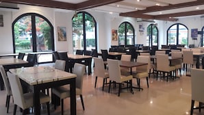 A spacious dining area with multiple tables and chairs, large arched windows, and artwork on the walls. - Grand Smile Wellness Resort (Pattaya)
