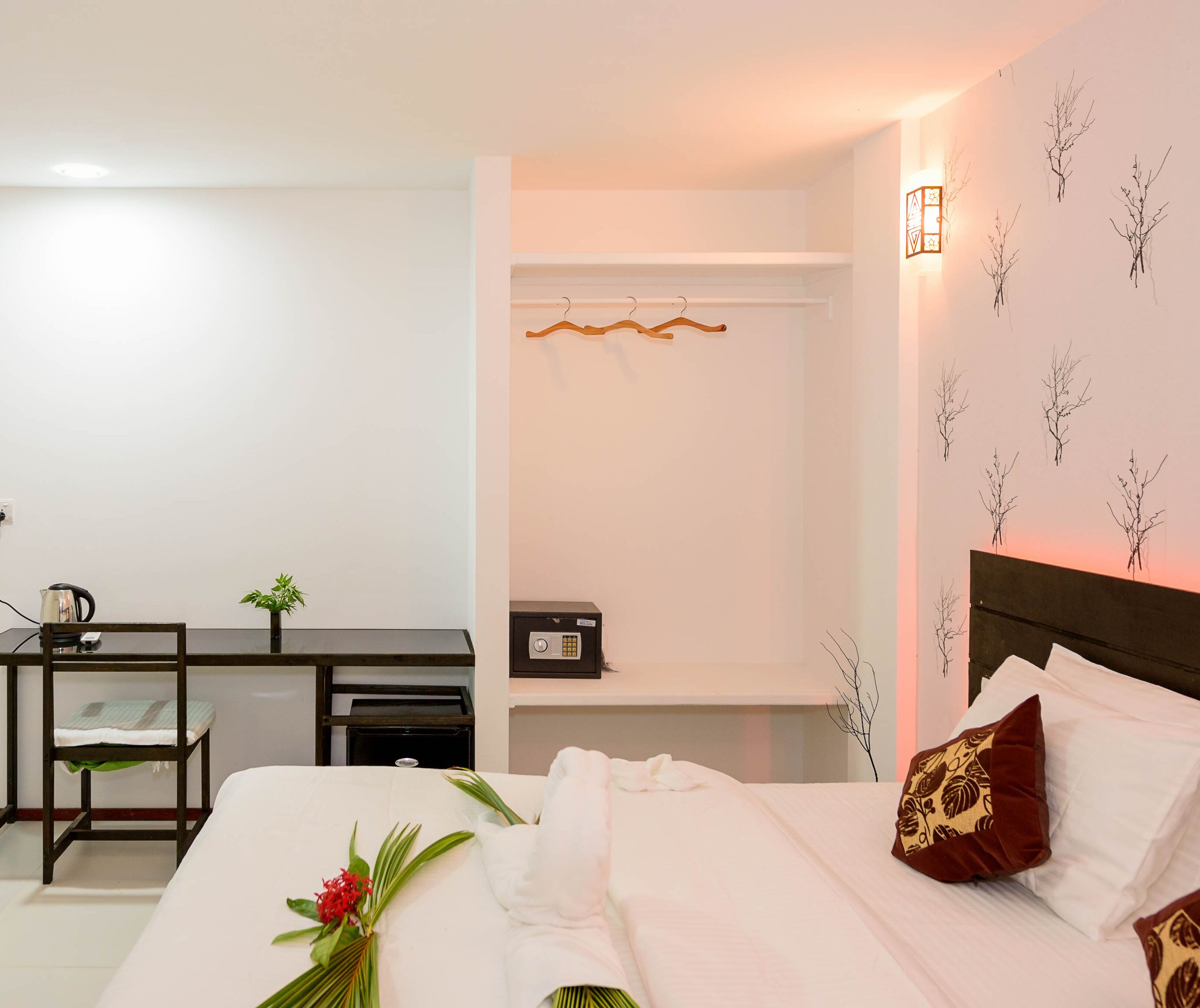 Una habitación de hotel con cama, escritorio, silla, mesita, planta y una lámpara fijada a la pared.