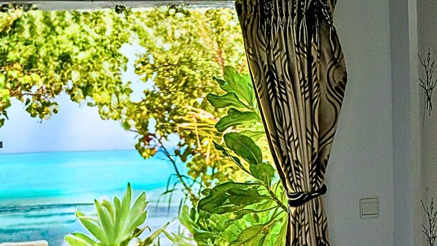 A hotel room with a bed, a small table, and a chair. There is a view of the ocean and trees outside the window. - Crown Beach Villas  Bikini Beach (Dhiffushi)