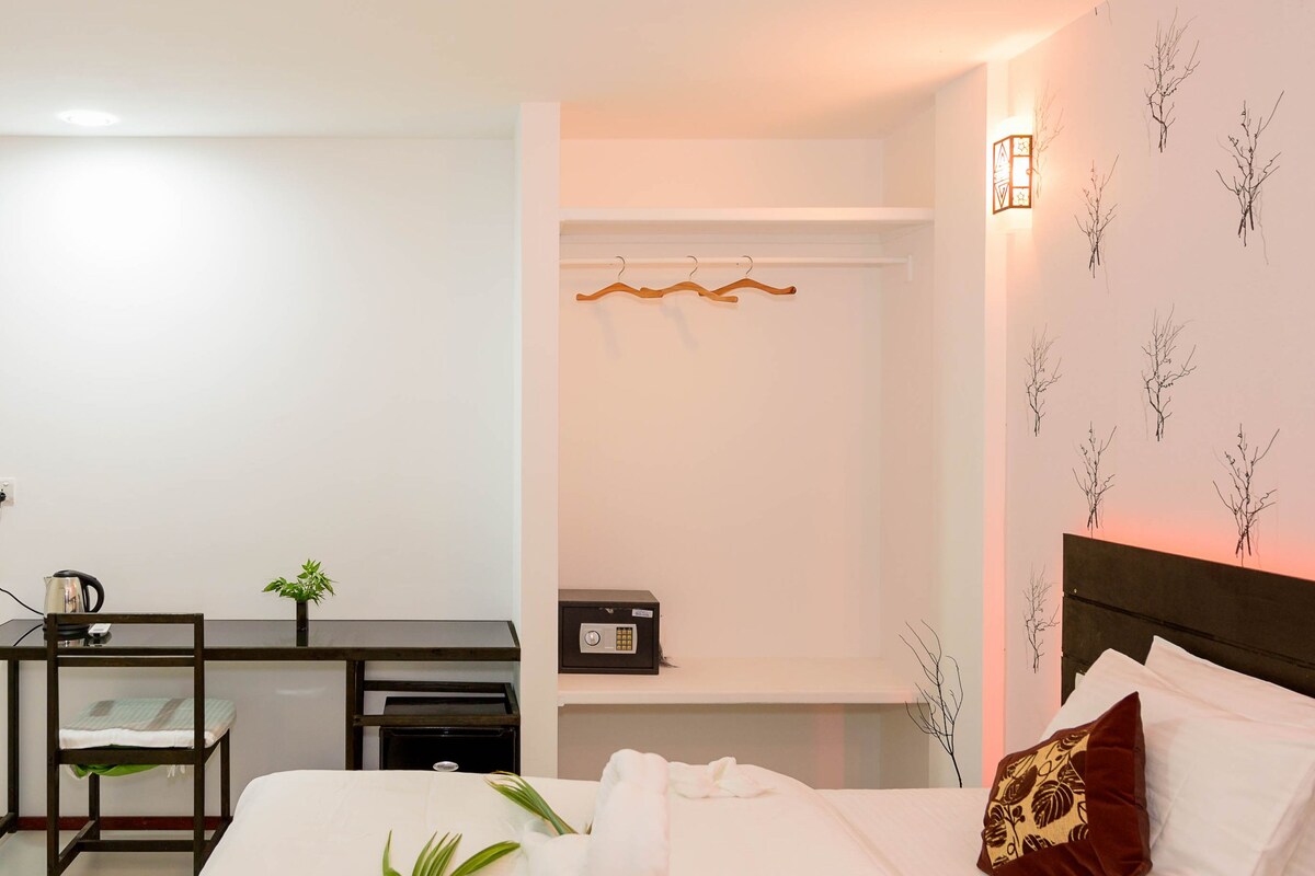 Une chambre d’hôtel comprenant un lit, un bureau, une chaise, une petite plante, un coffre-fort et des œuvres d’art murales.
